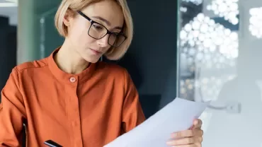 Frau mit blonden Haaren und Brille liest ein Blatt Papier