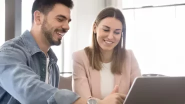 Zwei Personen sitzen an einem Tisch vor einem Laptop und lächeln. 