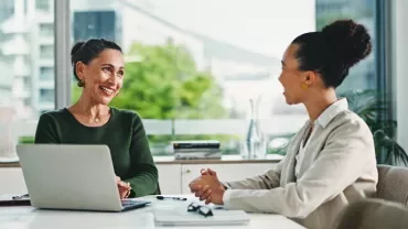 Zwei Frauen im Gespräch an einem Tisch, mit einem geöffneten Laptop zwischen ihnen. 