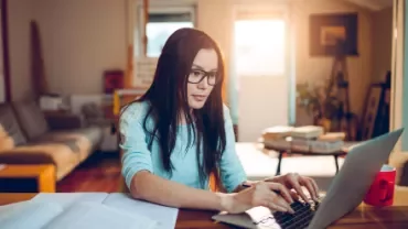 Frau mit Brille arbeitet an Laptop in gemütlichem Wohnzimmer.