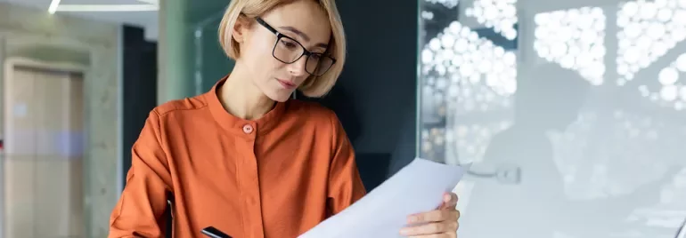 Frau mit blonden Haaren und Brille liest ein Blatt Papier