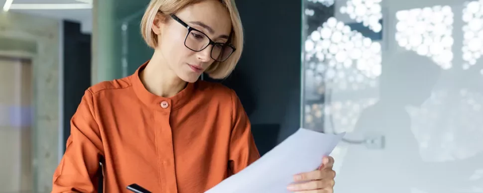 Frau mit blonden Haaren und Brille liest ein Blatt Papier
