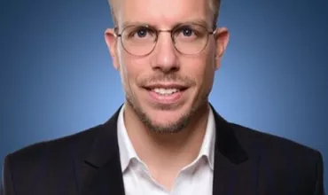 Mann im Anzug mit Brille vor blauem Hintergrund.